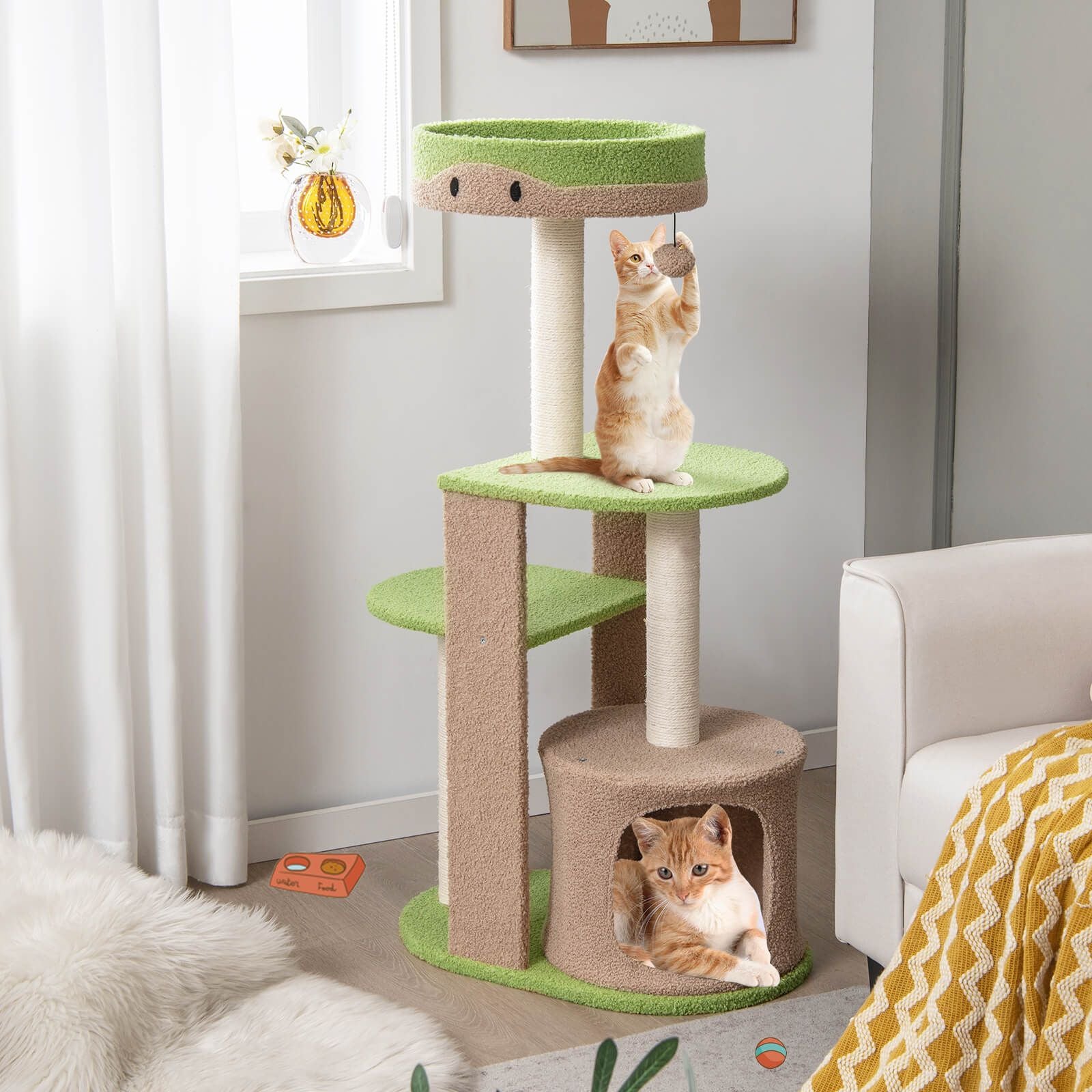 A multi-level cat tree with green and beige platforms, featuring two orange tabby cats playing. One cat is reaching up from a middle platform, while another sits in a cubby at the bottom. The tree is placed in a bright, modern living room setting with a window, decorative artwork, and a partial view of a white couch and yellow throw